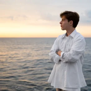 Person in a white shirt stands with arms crossed, looking out over a calm sea at sunset under a partly cloudy sky.