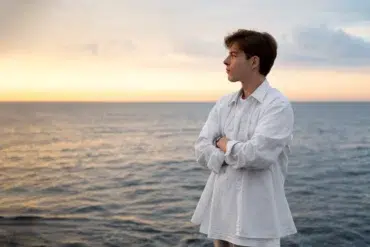 Person in a white shirt stands with arms crossed, looking out over a calm sea at sunset under a partly cloudy sky.