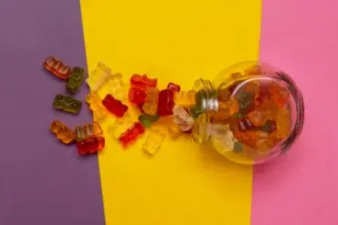 A glass jar spills colorful gummy bears onto a purple and yellow background, creating a playful and vibrant scene with assorted red, orange, and green candies.