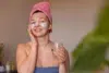 A woman with a facial mask and eye patches smiles warmly, holding a glass of water. She wears a pink towel turban and a blue wrap, suggesting relaxation.