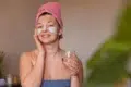 A woman with a facial mask and eye patches smiles warmly, holding a glass of water. She wears a pink towel turban and a blue wrap, suggesting relaxation.