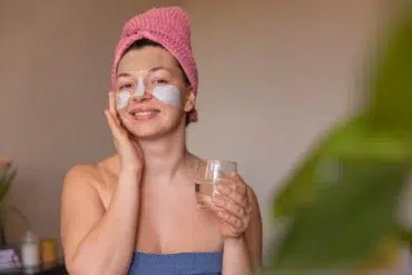 A woman with a facial mask and eye patches smiles warmly, holding a glass of water. She wears a pink towel turban and a blue wrap, suggesting relaxation.