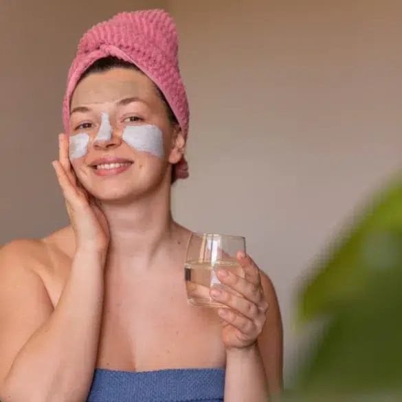 A woman with a facial mask and eye patches smiles warmly, holding a glass of water. She wears a pink towel turban and a blue wrap, suggesting relaxation.