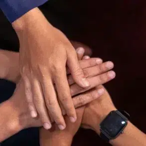 A group of four hands stacked together, symbolizing unity and teamwork. One wrist features a black smartwatch. The background is a dark color.