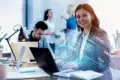 Smiling woman in a suit sits at a desk with a laptop, surrounded by holographic programming languages like PHP and Java. Colleagues converse in the background.