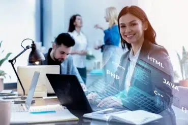 Smiling woman in a suit sits at a desk with a laptop, surrounded by holographic programming languages like PHP and Java. Colleagues converse in the background.