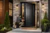 A worker kneeling on a porch installs a lock on a modern black door. The entrance features stone and wood accents, with symmetric potted plants.