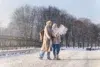 Two women in winter coats and hijabs stand on a snowy path, studying a map. Bare trees line the background under a clear sky, creating a serene scene.