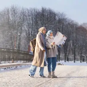 Two women in winter coats and hijabs stand on a snowy path, studying a map. Bare trees line the background under a clear sky, creating a serene scene.