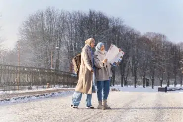 Two women in winter coats and hijabs stand on a snowy path, studying a map. Bare trees line the background under a clear sky, creating a serene scene.