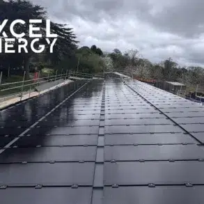A large array of solar panels installed on a flat rooftop under cloudy skies with trees and construction fencing nearby.