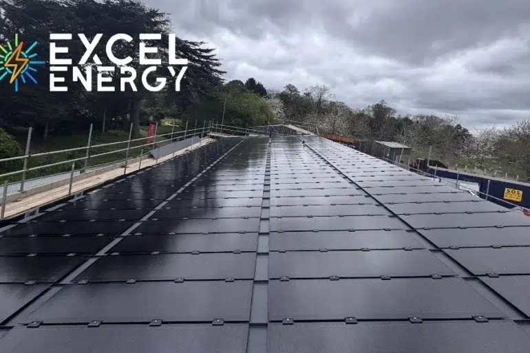 A large array of solar panels installed on a flat rooftop under cloudy skies with trees and construction fencing nearby.