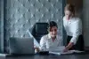 Two women in an office setting review documents at a desk with a laptop. Both wear white shirts, conveying a professional and focused atmosphere.