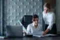 Two women in an office setting review documents at a desk with a laptop. Both wear white shirts, conveying a professional and focused atmosphere.
