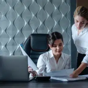 Two women in an office setting review documents at a desk with a laptop. Both wear white shirts, conveying a professional and focused atmosphere.