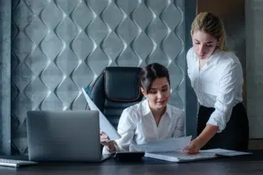 Two women in an office setting review documents at a desk with a laptop. Both wear white shirts, conveying a professional and focused atmosphere.