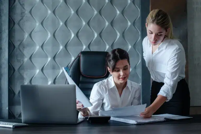 Two women in an office setting review documents at a desk with a laptop. Both wear white shirts, conveying a professional and focused atmosphere.