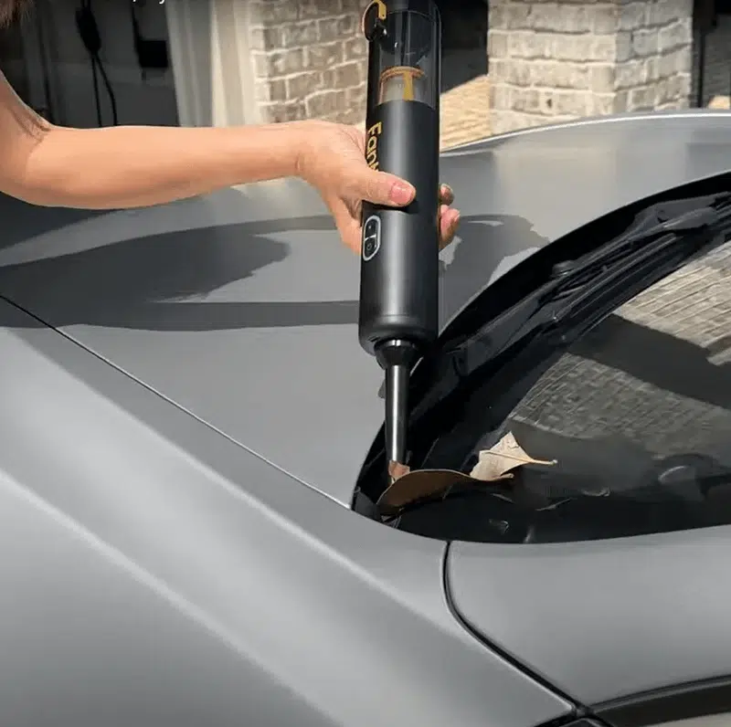 A person uses a black handheld vacuum to clean dry leaves from a car's windshield vent. The scene conveys practicality and tidiness.