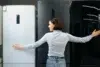 A woman in a light blue shirt and jeans embraces two refrigerators in a showroom, smiling as she compares a white and a dark model.