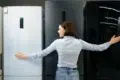 A woman in a light blue shirt and jeans embraces two refrigerators in a showroom, smiling as she compares a white and a dark model.