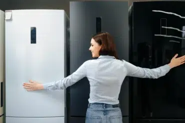 A woman in a light blue shirt and jeans embraces two refrigerators in a showroom, smiling as she compares a white and a dark model.