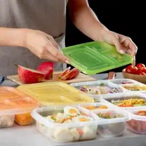 A person in an apron places a green lid on a meal prep container. Surrounding containers hold various foods, and a sliced apple, cherry tomatoes, and onions are nearby.