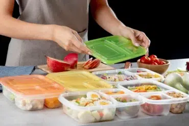 A person in an apron places a green lid on a meal prep container. Surrounding containers hold various foods, and a sliced apple, cherry tomatoes, and onions are nearby.