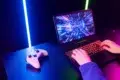 Person at a glowing laptop with a vibrant screen showing digital graphics. A gaming controller lies nearby on the table, creating an intense gaming vibe.