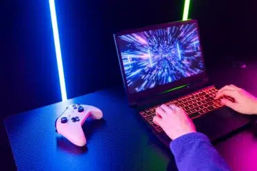 Person at a glowing laptop with a vibrant screen showing digital graphics. A gaming controller lies nearby on the table, creating an intense gaming vibe.