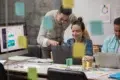 Three colleagues collaborate in a modern office, focused on laptops. Colorful sticky notes cover glass. Expressions are engaged and productive.