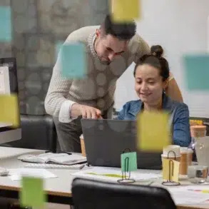 Three colleagues collaborate in a modern office, focused on laptops. Colorful sticky notes cover glass. Expressions are engaged and productive.