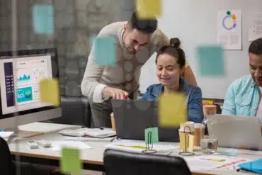 Three colleagues collaborate in a modern office, focused on laptops. Colorful sticky notes cover glass. Expressions are engaged and productive.