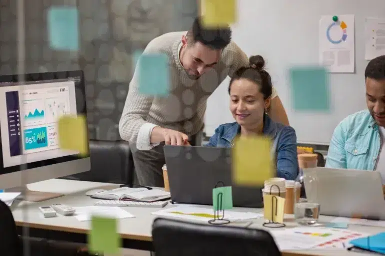 Three colleagues collaborate in a modern office, focused on laptops. Colorful sticky notes cover glass. Expressions are engaged and productive.