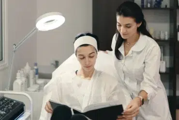 A woman in a spa chair reads a book, wearing a headband and robe, while another woman in a lab coat stands beside her, exuding a calm atmosphere.