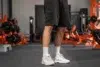 A person stands in a gym, wearing black shorts, white socks, and white sneakers. Gym equipment with orange accents is visible in the blurred background.