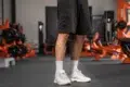 A person stands in a gym, wearing black shorts, white socks, and white sneakers. Gym equipment with orange accents is visible in the blurred background.