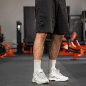A person stands in a gym, wearing black shorts, white socks, and white sneakers. Gym equipment with orange accents is visible in the blurred background.