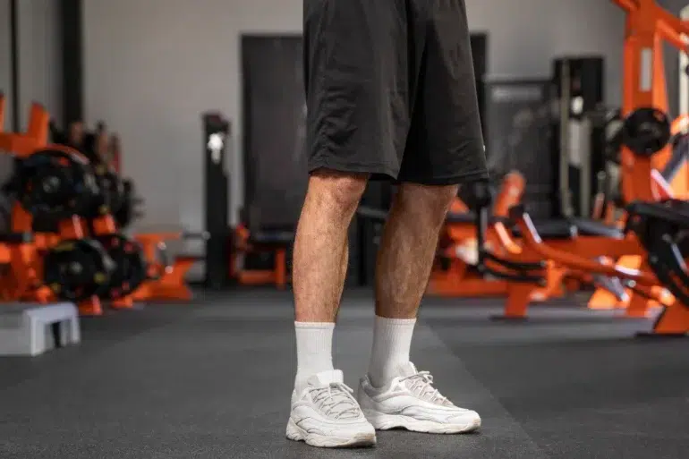 A person stands in a gym, wearing black shorts, white socks, and white sneakers. Gym equipment with orange accents is visible in the blurred background.