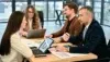 Four professionals sit around a table in a meeting room, discussing colorful charts on laptops and papers, conveying teamwork and focus.