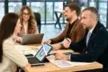 Four professionals sit around a table in a meeting room, discussing colorful charts on laptops and papers, conveying teamwork and focus.