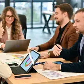 Four professionals sit around a table in a meeting room, discussing colorful charts on laptops and papers, conveying teamwork and focus.