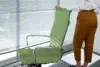 Person standing next to a green office chair in front of a window with closed blinds, wearing brown pants and a white shirt.
