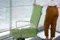 Person standing next to a green office chair in front of a window with closed blinds, wearing brown pants and a white shirt.