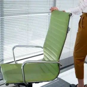 Person standing next to a green office chair in front of a window with closed blinds, wearing brown pants and a white shirt.