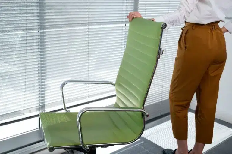 Person standing next to a green office chair in front of a window with closed blinds, wearing brown pants and a white shirt.