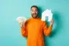 A joyful man in an orange sweater holds dollar bills and a white paper house cutout against a teal background, conveying financial success and homeownership.