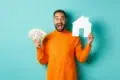 A joyful man in an orange sweater holds dollar bills and a white paper house cutout against a teal background, conveying financial success and homeownership.