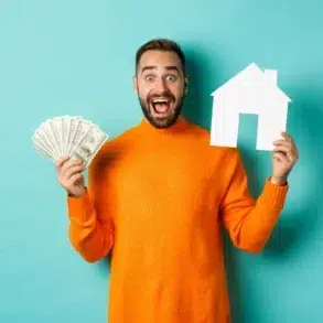 A joyful man in an orange sweater holds dollar bills and a white paper house cutout against a teal background, conveying financial success and homeownership.