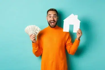 A joyful man in an orange sweater holds dollar bills and a white paper house cutout against a teal background, conveying financial success and homeownership.