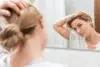 A woman in a white t-shirt uses a bathroom mirror to adjust her human hair topper for women.
